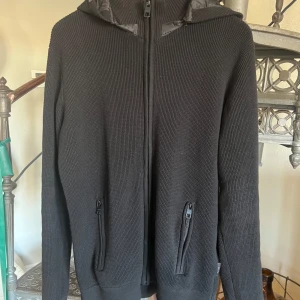 Svart cardigan från indicode - Snygg svart stickad jacka med dragkedja och huva. Perfekt för kyliga dagar. Jackan har långa ärmar och praktiska fickor med dragkedja framtill. Väldigt sällsynt då de är slutsålda överallt! Det är bara att köpa! Storlek L