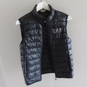 Svart dunväst från Emporio Armani - Snygg svart dunväst från Emporio Armani med dragkedja framtill. Västen har en stilren design med EA7-logga på bröstet och är perfekt för kyligare dagar. Den quiltade stilen ger ett modernt intryck och passar bra till olika outfits.