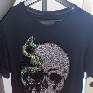 Svart t-shirt med dödskalle och orm från Philipp Plein - Snygg svart t-shirt från Philipp Plein med en stor glittrig dödskalle och en grön orm som slingrar sig runt motivet.  Kortärmad och i ett mjukt material.