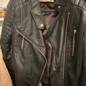 Svart kappa med guldknapp - Elegant svart kappa med en enkel guldknapp framtill. Kappan har en klassisk design med långa ärmar och två fickor. Perfekt för en stilren look.