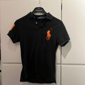 Svart kortärmad pikétröja från Polo Ralph Lauren - Snygg svart pikétröja från Polo Ralph Lauren med korta ärmar. Tröjan har en stor orange broderad logga på bröstet och en orange siffra på ärmen. 