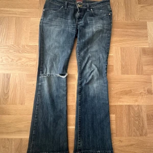 Blå bootcut jeans - Säljer ett par klassiska blå bootcut jeans med slitningar på knäet. De har en låg midja och femficksdesign med dragkedja och knapp framtill. Perfekta för en avslappnad stil.