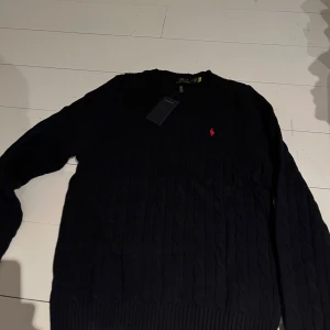 Svart kabelstickad tröja från Polo Ralph Lauren - Säljer en stilren svart kabelstickad tröja från Polo Ralph Lauren. Tröjan har en klassisk design med rund hals och det ikoniska röda logotypbroderiet på bröstet. Perfekt för en elegant och avslappnad look.