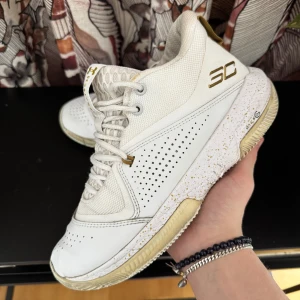 Träningskor - Snygga vita sneakers från Under Armour med guldiga detaljer och perforerad design. Skorna har snörning och en bekväm passform, perfekt för både sport och vardag. Den vita färgen kombineras med subtila guldinslag för en stilren look.