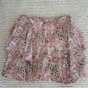 Rosa blommig kjol med volang - Säljer en söt rosa kjol med blommigt mönster och volangdetaljer. Perfekt för en somrig look. Kjolen har en omlott-design som ger en fin passform. Passar bra till både vardag och festliga tillfällen.