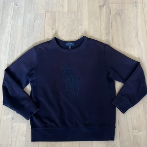 Mörkblå tröja från Ralph Lauren - Säljer en stilren mörkblå tröja från Ralph Lauren med ett diskret broderat motiv på framsidan. Tröjan har långa ärmar och en klassisk rund halsringning. Perfekt för en avslappnad och snygg look.