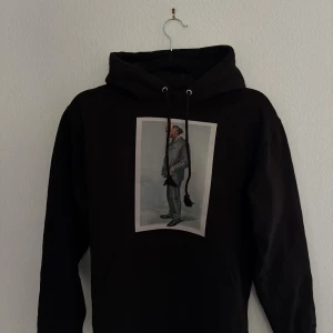 Svart hoodie från The Cool Elephant - Svart hoodie med stor huva och snörning från The Cool Elephant. Framsidan har ett tryck med en tecknad man i kostym, och på ryggen finns ett vitt elefantlogotyp-tryck. Tröjan har en klassisk känguruficka.