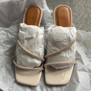 Beigea klackar från NAKD - Säljer ett par stilrena sandaler från NA-KD i beige med transparenta remmar och glittriga strassdetaljer. ANVÄNDA 1 GÅNG. Nypris 599