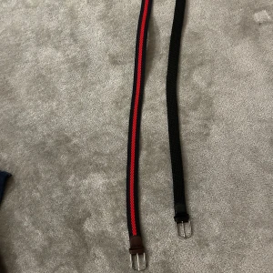 Två flätade bälten från John Henric - Säljer två snygga flätade bälten från John Henric. Ett är helsvart med svarta skinn-detaljer, det andra har svart bas med en röd rand i mitten och bruna skinn-detaljer. Båda har silvriga spännen och är märkta med storlek 100.