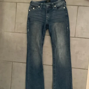 True Religion Becca Bootcut Jeans - Snygga blå bootcut jeans från True Religion, modell Becca Mid Rise. Jeansen har glittriga detaljer på bakfickorna och stora kristallknappar. Klassisk femficksmodell i stretchig denim med cool tvätt och markerade sömmar. Använda fåtal gånger. Priset kan diskuteras💗