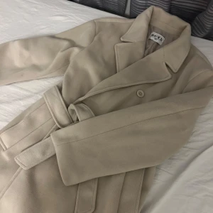 Beige kappa med bälte från NA-KD - Snygg beige kappa från NA-KD i storlek 36. Klassisk design med bred krage, knappar framtill och matchande bälte i midjan. Jackan har långa ärmar och är tillverkad i ett mjukt material. Jag är 163 och jackan går ner till exakt under mitt knäskål