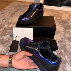 Blå metallic sneakers från Giuseppe Zanotti - Snygga sneakers från Giuseppe Zanotti i blå metallic med reptilmönster och svarta detaljer. Skorna har svarta snören, dragkedjor på sidorna och svart sula. Lyxig och unik design som sticker ut, perfekt för dig som gillar statement pieces.