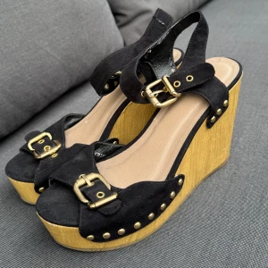 Svarta platåsandaler med träklack - Snygga svarta sandaler med bred kilklack träklack och platåsula. Ovandel i mocka med guldfärgade spännen och nitar som ger en cool retrovibe. Öppen tå och justerbara remmar runt vristen. Perfekta för dig som vill sticka ut med höga klackar och statement-detaljer. Använda så skavanker förekommer, vg se bilder.. Storlek 38. Booho look