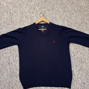 Mörkblå v-ringad tröja Polo Ralph Lauren - Snygg mörkblå v-ringad tröja från Polo Ralph Lauren i storlek L men känns som M. Tröjan har ett rött broderat Polo-logo på bröstet och är tillverkad i mjuk bomull. Långärmad modell med ribbade muddar vid ärmslut och nederkant. Perfekt för dig som gillar klassisk stil.