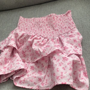 Rosa blommig volangkjol - Supersöt kort kjol i ljusrosa med små rosa blommor. Kjolen har dubbla volanger som ger en flowy känsla och är perfekt för varma dagar. Materialet känns lätt och luftigt, perfekt för sommarens alla äventyr.