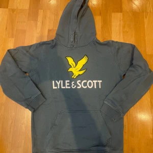  hoodie från Lyle & Scott - Snygg grön hoodie från Lyle & Scott med stor gul logga och vit text på bröstet. Tröjan har huva, magficka och ribbade muddar vid ärmslut och nederkant. Perfekt för en avslappnad och cool stil. Använd ett fåtal gånger.