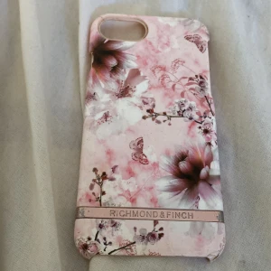 Richmond & Finch blommigt mobilskal rosa - Snyggt mobilskal från Richmond & Finch med blommigt mönster i rosa och vita toner. Skalet har en ljusrosa bakgrund med detaljerade blommor och fjärilar, samt en metallisk rand med loggan. Passar perfekt för dig som vill ha en personlig och trendig look på mobilen.