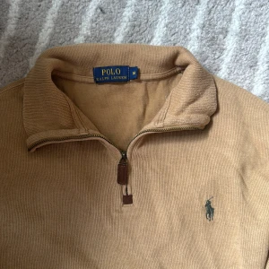 Beige half zip tröja från Polo Ralph Lauren - Snygg beige långärmad tröja från Polo Ralph Lauren med half zip och klassisk broderad logga på bröstet. Tillverkad i mjuk bomull med ribbad struktur och hög krage. 