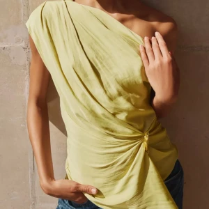 Gul oneshoulder topp med drapering - Snygg ljusgul oneshoulder topp med draperad design och cool detalj med knyt i sidan. Tunt, flowigt material som ger en lyxig känsla och passar perfekt till jeans eller kostymbyxor. Asymmetrisk nederkant för extra edge.