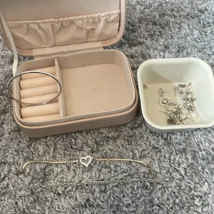 Mix av silversmycken med hjärtarmband - Säljer ett paket med silvriga smycken: ett stelt armband med glittrande stenar, ett armband med hjärtdetalj och strass, en tunn kedja samt flera olika örhängen i silverfärg. Perfekt för dig som gillar att mixa och matcha accessoarer. Medföljer smyckeslådan, går att köpa separat