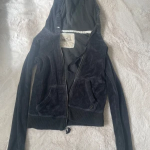 Vintage Hollister zip up - Svart velourjacka från Hollister med huva och dragkedja framtill. Jackan har två fickor, ribbade muddar och en liten metallogo vid ena fickan. Perfekt för en chill och avslappnad stil.