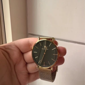 Svart och guld klocka från Black Oak - Stilren klocka från Black Oak med svart urtavla och tunna guldiga markeringar. Boetten är rund och i guldton, och armbandet är ett snyggt meshband i metall med guldfinish. Perfekt för dig som gillar minimalistisk design och vill ha något som sticker ut.