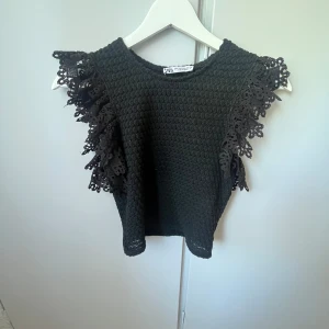Svart topp med spetsvolang från Zara - Supersnygg svart topp från Zara med coolt material och breda spetsvolanger på axlarna. Toppen är ärmlös och har en croppad passform som ger en trendig vibe. Perfekt att styla med höga jeans eller kjol för en extra touch. Står S men är en xs typ- och är även lite croppad💕💕