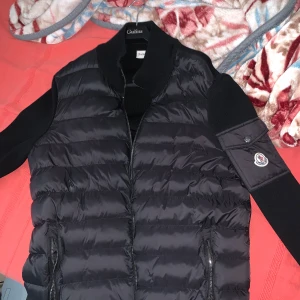 Svart dunväst från Moncler, storlek L - Säljer en svart dunväst från Moncler i storlek L. Västen har quiltad design, dragkedja framtill och två fickor med dragkedja. Ärmar och sidor är i stickat material och vänster ärm har Moncler-logga på en ficka. Perfekt för lager-på-lager och snygg streetstil.