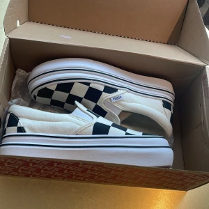 Vans Checkerboard Slip-On Sneakers - Klassiska Vans slip-on sneakers med svartvitt rutigt mönster. Skorna har vit sula med svart rand och den ikoniska röda undersulan. Ovandelen är i canvas och insidan har en bekväm dämpad innersula. Perfekta för en avslappnad och cool streetstil.