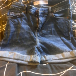 Blå jeansshorts från Amisu, strl 36 - Snygga blå jeansshorts från Amisu i storlek 36. Klassisk femficksmodell med uppvikta benslut och dragkedja framtill. Perfekta för sommaren och lätta att matcha med allt. Sköna och stilrena shorts för en avslappnad look.
