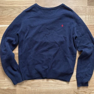 Mörkblå stickad tröja Morris Gents - Mörkblå stickad tröja från Morris Gents i storlek S. Klassisk modell med rund halsringning, ribbade muddar och en liten röd broderad detalj på bröstet. Perfekt för lager-på-lager och snygg till jeans eller chinos.