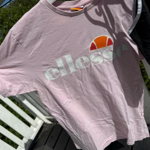 Ljusrosa Ellesse t-shirt med logga - Säljer en ljusrosa t-shirt från Ellesse med stor vit logga och orange detalj framtill. Klassisk rund hals och korta ärmar, perfekt för en chill och sportig look. Tillverkad i mjuk bomull som känns skön mot huden.