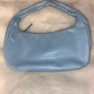 Ljusblå axelväska i syntet - Säljer en stilren ljusblå axelväska med mjuk, rundad form och enkel design. Väskan har ett kort handtag och stängs med dragkedja upptill. Perfekt för dig som vill ha en clean och trendig look.