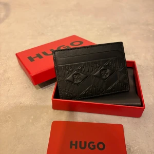 Svart plånbok från Hugo i skinn - Snygg svart plånbok från Hugo i äkta skinn med präglat geometriskt mönster och logga. Slimmad design med plats för kort, perfekt att ha i fickan. Kommer i original röd Hugo-box, riktigt stilren och modern vibe. Helt nyskick! 
