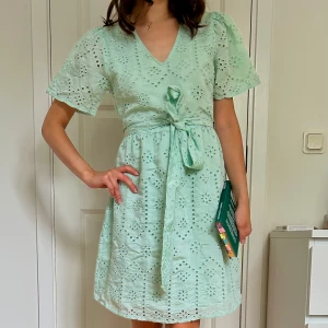 Ljusgrön broderad Vila klänning ny med prislapp. - Säljer en ljusgrön kort klänning med v-ringning och knyt i midjan. Klänningen har broderade detaljer och ett fint hålmönster över hela tyget. Puffiga korta ärmar ger en söt och somrig vibe. Perfekt för dig som gillar romantisk och feminin stil💚