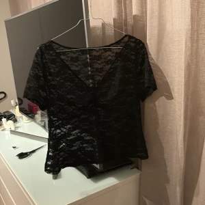 Svart spetsblus med v-ringning - Säljer en svart blus i genombruten spets med v-ringning och korta ärmar. Blusen är lätt och luftig, med snyggt mönster i spetsen. Storlek M 