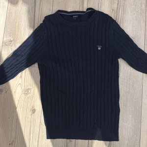  Kabelstickad tröja från GANT - Snygg mörkblå stickad tröja från GANT med diskret logga på bröstet. Tröjan har ett klassiskt kabelstickat mönster, rund halsringning och långa ärmar. Perfekt för dig som gillar en stilren och tidlös look.