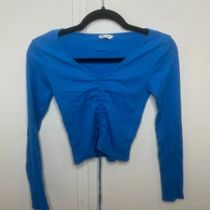 Blå croppad topp med snörning - Snygg blå långärmad topp från Cubus i ribbad bomull. Croppad modell med v-ringning och cool snörning framtill som ger en unik look. Perfekt för dig som gillar att sticka ut med färg och detaljer. Passar dig som vill ha en slim fit och trendig stil.