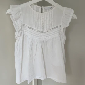 Vit blus med volang  - Supersöt vit blus från Name It med volangärmar och fina broderade detaljer framtill. Blusen har rund hals med knapp i nacken! Storlek 134-140 men ungefär som en xxs! 