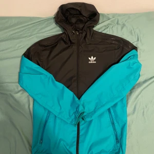 Adidas svart och turkos vindjacka XS - Snygg vindjacka från Adidas i svart och turkos med huva och dragkedja. Jackan har klassiska tre ränder på ena ärmen, två fickor med dragkedja och Adidas-logga på bröstet. Perfekt för dig som gillar sportig och street-inspirerad stil.