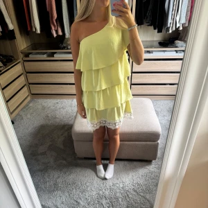 Gul klänning med volanger - Supersöt ljusgul kortklänning med omlott-design och flera lager volanger. Klänningen är oneshoulder och har en vit spetskant med blommönster längst ner. Perfekt för sommarens alla tillfällen, riktigt fräsch och ungdomlig vibe.
