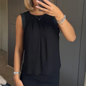 Svart ärmlös blus - Säljer en svart ärmlös blus med snygga veck framtill och skinndetaljer vid axlarna som ger en stilren och chic look. Blusen har rund halsringning och är tillverkad i ett lätt material som faller snyggt.