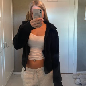 Svart teddyjacka från revolutionrace  - Mysig svart teddyjacka med dragkedja framtill, huva med snörning och ribbade muddar. Jackan är fluffig och har en enkel, stilren look som passar perfekt till chill outfits. Broderad logga på bröstet och två fickor med dragkedja.