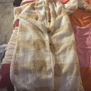 Lång teddyjacka i beige och vitt - Supermysig lång teddyjacka i beige och vitt med stora knappar framtill. Jackan har ett rutigt mönster och är gjord i fluffigt teddy-material som håller dig varm. Perfekt för kalla dagar och har en oversized vibe. Snygg att slänga över vilken outfit som helst.
