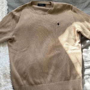 Beige stickad tröja Polo Ralph Lauren - Snygg beige stickad tröja från Polo Ralph Lauren med klassisk rund hals och broderad logga på bröstet. Tröjan har ribbade muddar vid hals, ärmslut och nederkant. Perfekt för dig som gillar stilrena och tidlösa plagg.