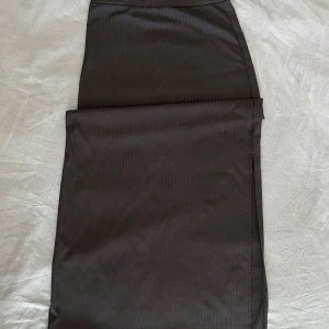 Svart ribbad långkjol från SHEIN - Svart långkjol från SHEIN i storlek S. Kjolen har en ribbad struktur och är figurnära med slits på sidan för extra stilpoäng. Perfekt för dig som gillar minimalistisk och clean look. Materialet är mjukt och stretchigt syntet.