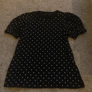 Svart prickig topp från Mywear Woman - Svart topp från Mywear Woman i storlek S med vita prickar över hela tyget. Modellen har korta puffärmar och rund halsringning. Perfekt för dig som gillar klassiska mönster med en söt twist.