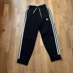 Svarta Adidas mjukisbyxor med vita ränder - Svarta mjukisbyxor från Adidas med klassiska vita ränder längs sidorna och Adidas-logga på låret. Byxorna har också vita snören.Tillverkade i mjukt material, perfekta för chill eller träning.Byxorna är rätt så anndvända,ena sidan av snörerna är en knut och de är lite noppriga men annars i bra skick!
