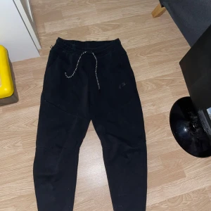 Svarta Nike tech byxor - Svarta mjukisbyxor från Nike med dragsko i midjan och diskret logga på benet. Byxorna har en relaxed fit och är tillverkade i mjukt bomullsmaterial, perfekta för chill eller träning. Snygga detaljer med kontrastfärgad snörning. Jag har använt dom 1 gång