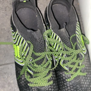 Svarta och gröna fotbollsskor från Fila - Snygga fotbollsskor från Fila i svart med gröna detaljer och snörning. Skorna har mönstrad ovandel med vita och gröna linjer samt gröna dobbar undertill för bra grepp på planen. Perfekta för dig som vill sticka ut på matchen!